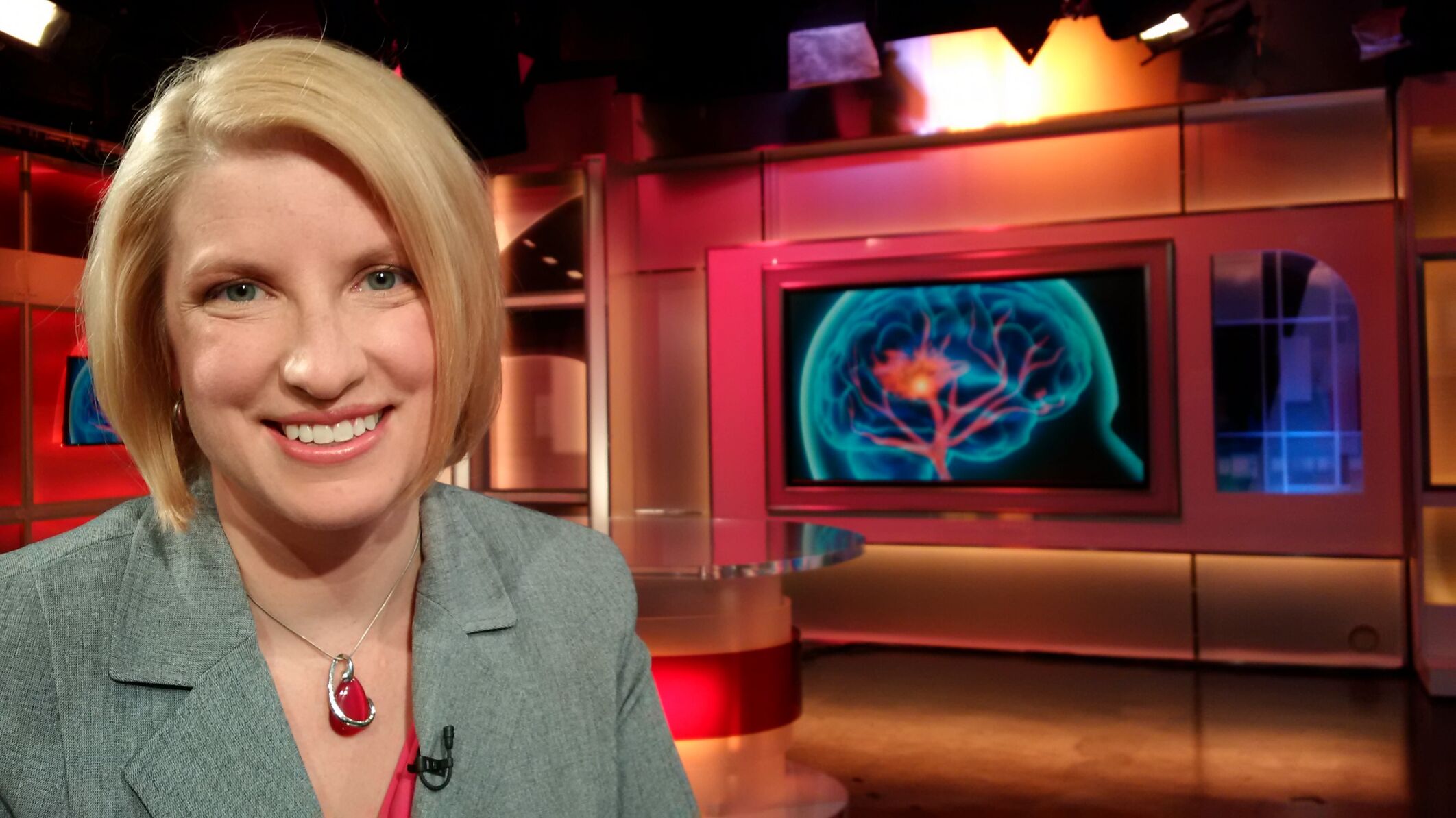 A female smiling at the camera on a filming set with a television showing an image of the brain during stroke